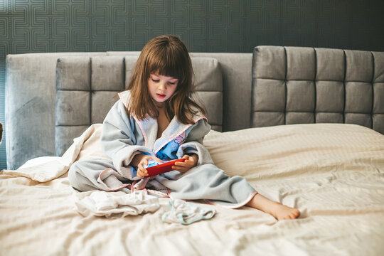 A young girl in a bathrobe is immersed in her smartphone, her attention fully captured by its screen. Concept: kids and smartphones