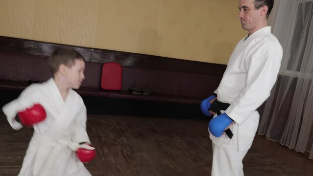 A trainer with a black belt and blue pads on his hands ties a white belt to a student and trains strikes and defense techniques with him