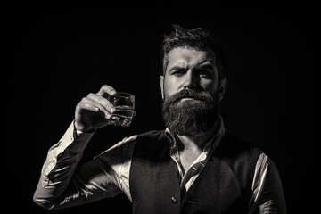 Man with beard holds glass with alcohol. Barman or degustator with curious face holds glass of cognac. Bearded man wearing suit and drinking whiskey, brandy, cognac. Black and white