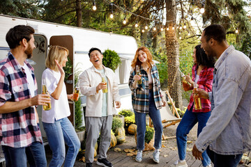 A cheerful group of friends rest in nature near a tourist trailer. Mixed-gender and multinational company of friends toasting, dancing, drinking drinks, have fun.