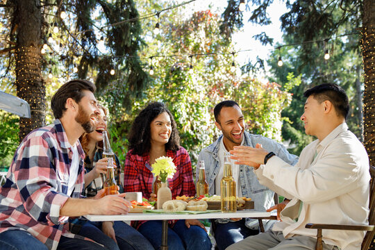 A cheerful multinational company is having fun on a picnic. Multisex friends sitting at the table outdoors celebrating eating and drinking drinks.
