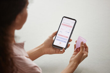 Over shoulder closeup of young woman holding smartphone with calendar app on screen and silicon menstrual cup - 787129658