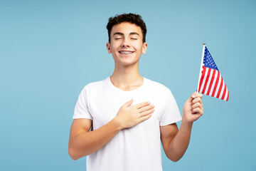 Smiling patriot young boy teenager holding American flag with eyes closed holding hand over heart