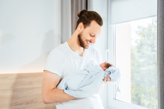 Proud young father admiring and calming his little newborn baby son at hospital room after labor.