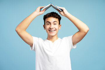 Attractive happy teenage boy with braces wearing casual white t shirt holding book above head