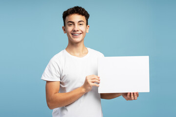 Happy smiling boy, teenager with braces holding white blank screen, copy space, looking at camera