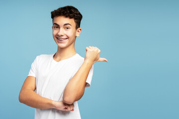 Happy teenage boy with braces showing finger on copy space standing isolated on blue background