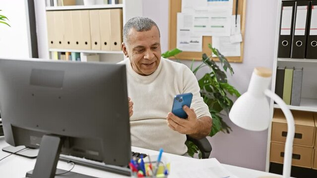 Mature hispanic man gives thumbs-up while using smartphone in an office with computer and binders.