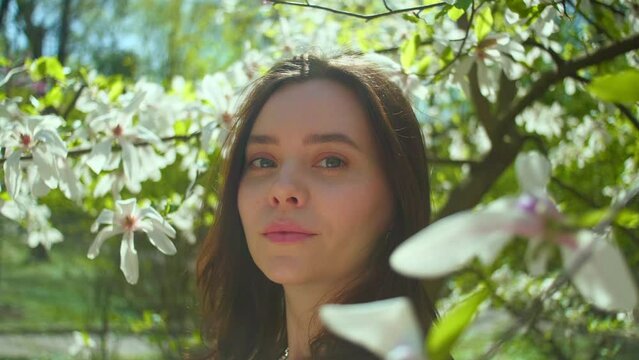 Beautiful woman smiling looks at camera. Girl in sunlight surrounded by soft haze of magnolia blossom petals, relaxed female in garden. Cinematic slow motion shot