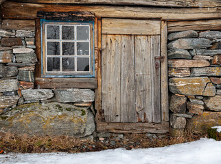 Weathered wood cabin door and window set in stone foundation amidst traces of snow.