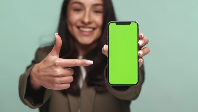 Cheerful young woman pointing at her phone with a mockup green screen for easy replacement, perfect for app or website promotion.