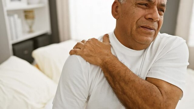 A smiling middle-aged hispanic man in a white shirt massages his shoulder, expressing relief in a bright bedroom setting. - Powered by Adobe