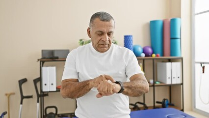A mature hispanic man checks his smartwatch in a rehabilitation clinic, highlighting healthcare technology in an indoor setting.