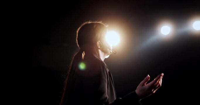 Speech of female speaker at business conference or training presentation. Back view at confident woman standing on stage in hall spotlights, talking motivating education