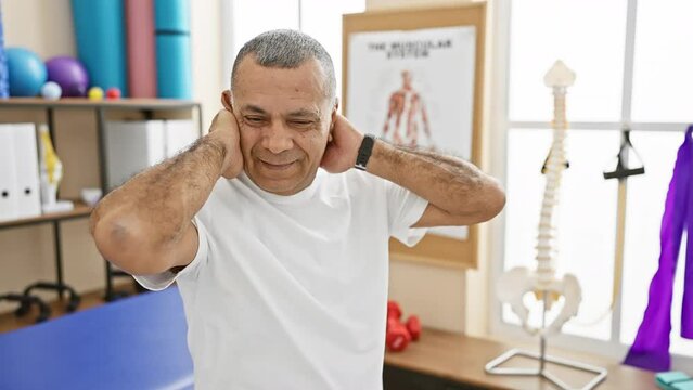 Middle-aged hispanic man stretching neck muscles indoors at a physical therapy clinic, showing discomfort and rehabilitation exercises.