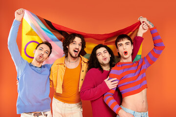 four joyous fashionable gay men in vibrant attires holding rainbow flag in front of camera, pride