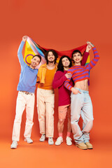 four joyful fashionable gay men in vibrant attires holding rainbow flag in front of camera, pride