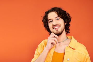 jolly appealing gay man with dark hair and vibrant makeup posing on orange backdrop, pride month