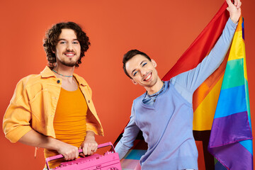 two contented handsome gay friends posing with tape recorder and rainbow flag on orange backdrop