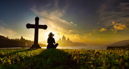 Silhouette of a young woman sitting on the grass praying in front of a cross at sunset and beautiful orange colored sky
