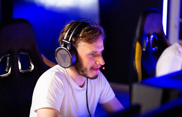 Adult bristly gamer in headset playing video game at computer club, cyber sport tournament. E-sport championship. Player in headphones communicates with others over mic. Multiplayer online competition - 787160849