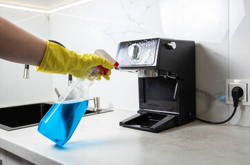 The girl holds in her hand an anti-grease spray for cleaning the kitchen from grease and dirt. Washing and cleaning a coffee machine and kitchen appliances against a white wall. Copy space for text