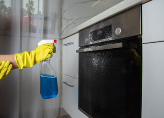 A girl holds a blue spray in her hand against the background of a modern electric oven in the...