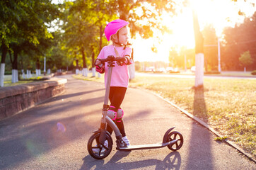 a beautiful girl in a pink helmet and protective equipment stands with a scooter on an alley in the city against the backdrop of a sunny sunset in summer. - 787161433
