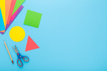 Colorful geometric shapes, scissors, pencil and application paper on light blue table background. Pastel color. Closeup. Child made different forms for learning. Empty place for text. Top down view. - 787164677