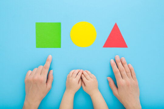 Young mother finger showing colorful geometric paper shapes to baby. Hands together on pastel blue table background. Time to learning. Infant development. Closeup. Point of view shot. Top down view.
