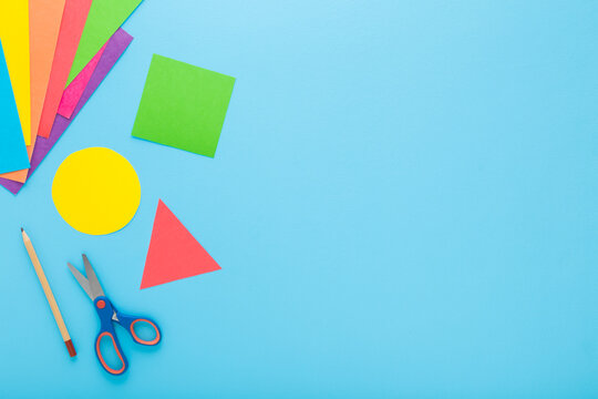 Colorful geometric shapes, scissors, pencil and application paper on light blue table background. Pastel color. Closeup. Child made different forms for learning. Empty place for text. Top down view.