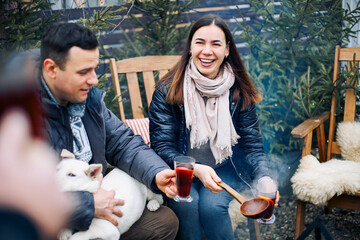 Winter bonfire party. Joyful young couple with dog drinking hot mulled wine in company of friends