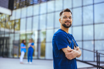 A male nurse is standing outdoors wearing scrubs and a stethoscope. He is smiling and looking at the camera. Portrait of male nurse. Portrait of mid adult nurse man at hospital - 787172008