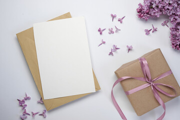 Blank greeting card mockup with gift box and lilac flowers on a white background. Flat lay card...