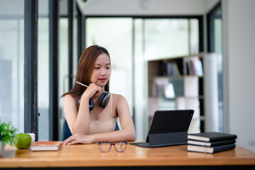 Smiling young woman with headphones sitting at a table with a laptop, enjoying music during study time at home.