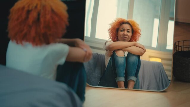 Young woman displeased with her reflection, sitting on the floor, insecurity