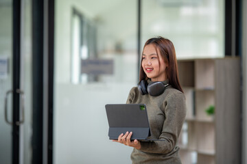 Happy young Asian woman holding a tablet with headphones around her neck in a bright, modern office.