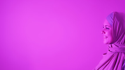 Smiling profile. Islamic fashion. Happy attractive woman in elegant hijab outfit in good mood on pink neon light copy space background.