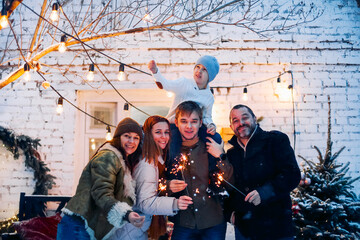 Portrait of big happy family with child standing in snow-covered house yard holding lit sparklers...