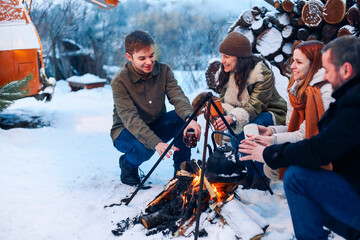 Group of friends gathering around bonfire in backyard, drinking tea and warming hands. Two happy couples relaxing and enjoying winter season while sitting around fire. Outdoor winter entertaining