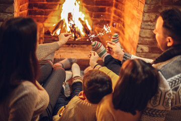 Happy young family parents with two small kids gathering around brick fireplace in cozy and warm country house during Christmas holidays.