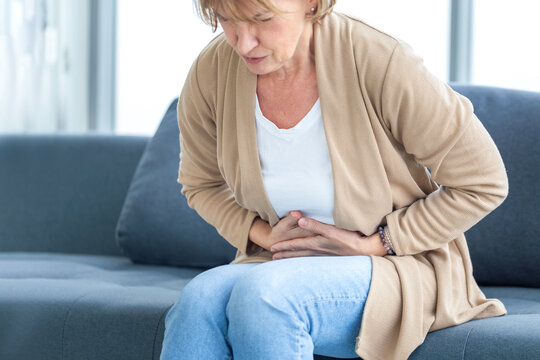 Unhappy woman stomach ache, mature woman with stomach pain feeling unwell sitting in living room