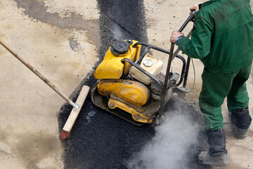 A road service worker compacts asphalt with a mechanical compactor. - obrazy, fototapety, plakaty