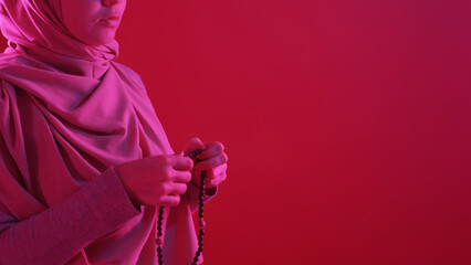 Islamic praying. Traditional tasbih. Unrecognizable woman with wooden rosary beads in vibrant pink neon light on red empty space background.