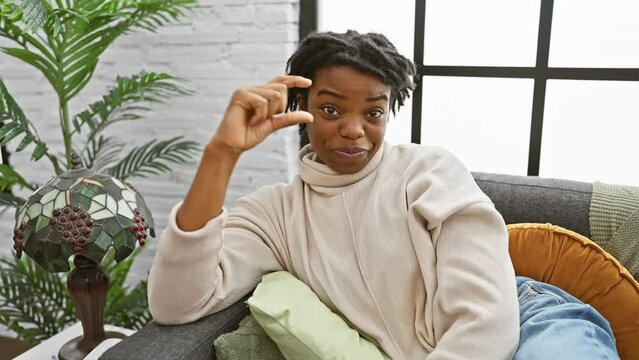 Joyful young black woman with dreadlocks, relaxing on a cozy sofa at home, confidently showing small size sign, gesturing a measure concept with her fingers.