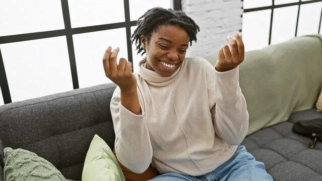 Sneaky yet triumphant, young black woman with dreadlocks, comfortably relaxing on a cozy sofa, doing a sly money gesture with her hands. home-based millionaire displaying her salary payment success.