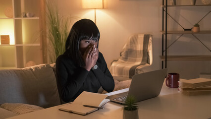 Shocked news. Excited woman. Online connection. Happy female student looking laptop covering mouth with hands in evening room interior.