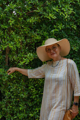 An elderly woman in a large hat and glasses stands next to a green bush, placing her hand on it, with a small handbag weighing on her shoulder.