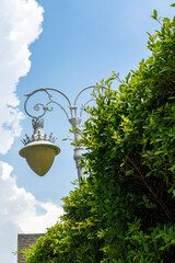 Against the background of a blue sky with white clouds, a night light lantern hangs on a pole. A green bush grows next to the lantern.