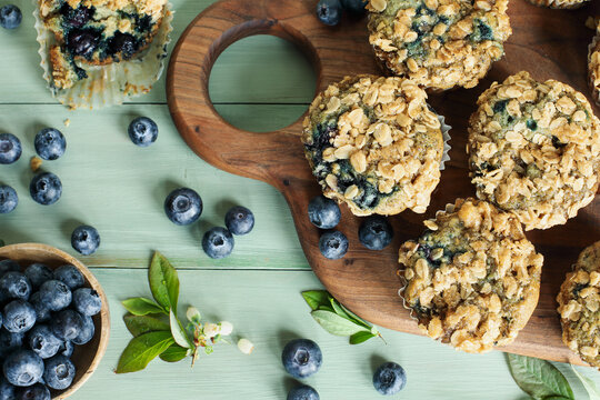 Fresh blueberry muffins with oat streusel topping and raw blueberries spilling from a wooden spoon. Table top view with focus on muffin tops and blurred background.